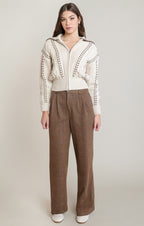 A model stands against a light beige studio background wearing a cream-colored zip-up knit cardigan with brown braided trim details and brown wide-leg trousers.