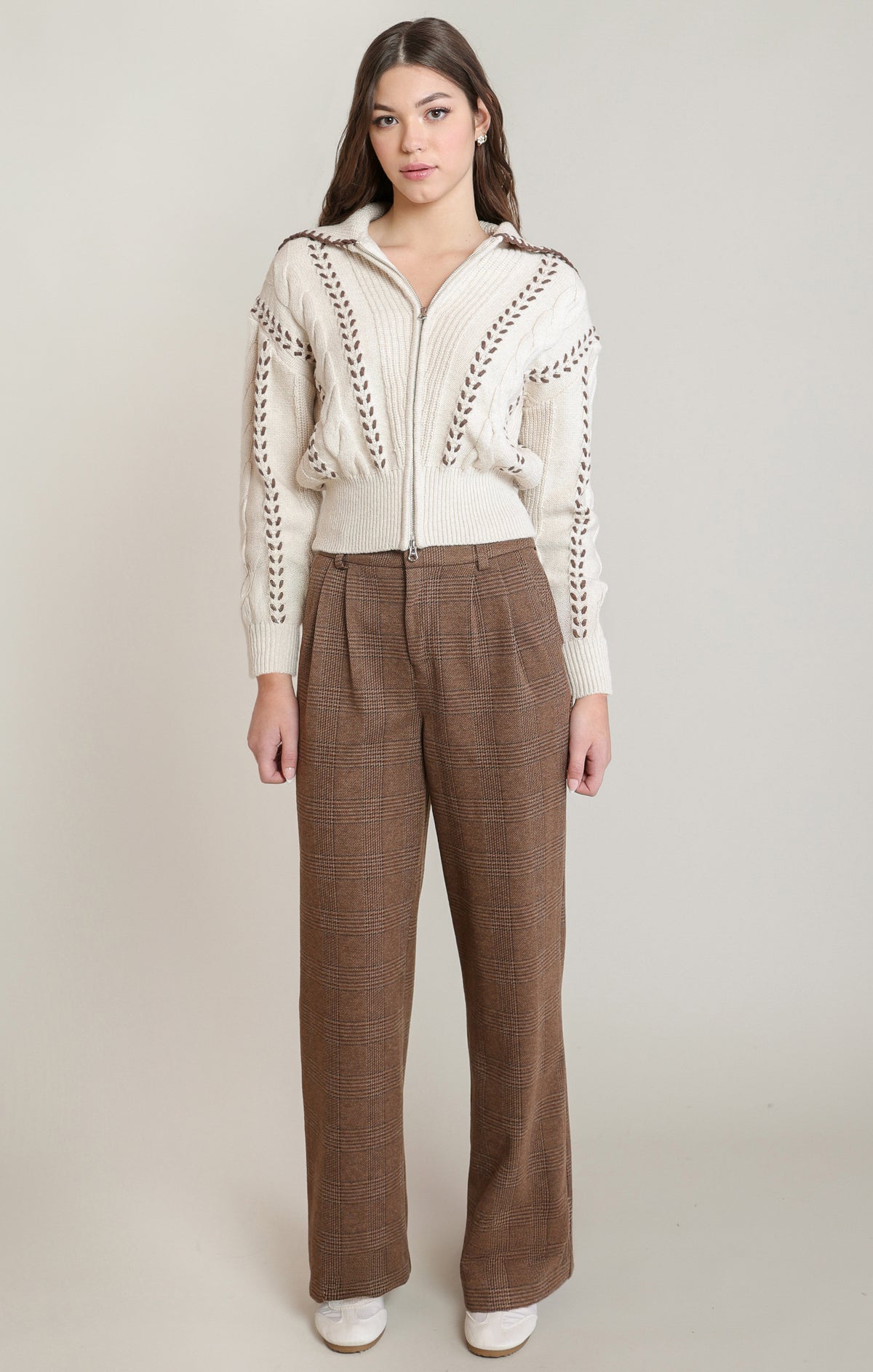 A model stands against a light beige studio background wearing a cream-colored zip-up knit cardigan with brown braided trim details and brown wide-leg trousers.