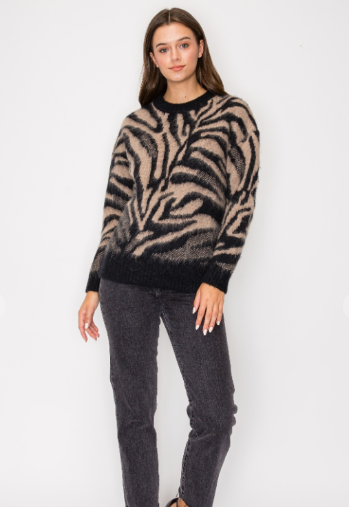 A woman modeling a fuzzy knit sweater with a beige and black zebra-like pattern, paired with dark gray jeans, against a plain white background.