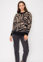 A woman modeling a fuzzy knit sweater with a beige and black zebra-like pattern, paired with dark gray jeans, against a plain white background.