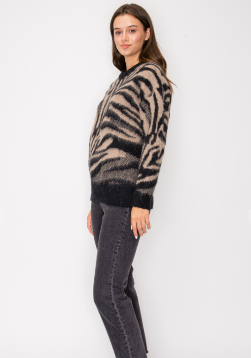 A woman modeling a fuzzy knit sweater with a black and beige zebra stripe pattern, paired with dark gray jeans, against a plain white studio background.