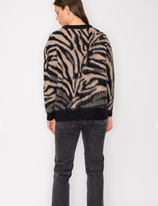 A woman viewed from the back wearing a fuzzy knit sweater with a zebra-like animal pattern in beige and black, paired with dark gray jeans, against a plain light gray studio background.