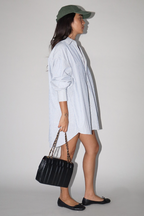 Person wearing a light blue shirt, dark green cap, and black flats holding a black handbag against a plain background