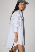 Woman wearing a long blue and white striped shirt dress, holding a bag against a plain background