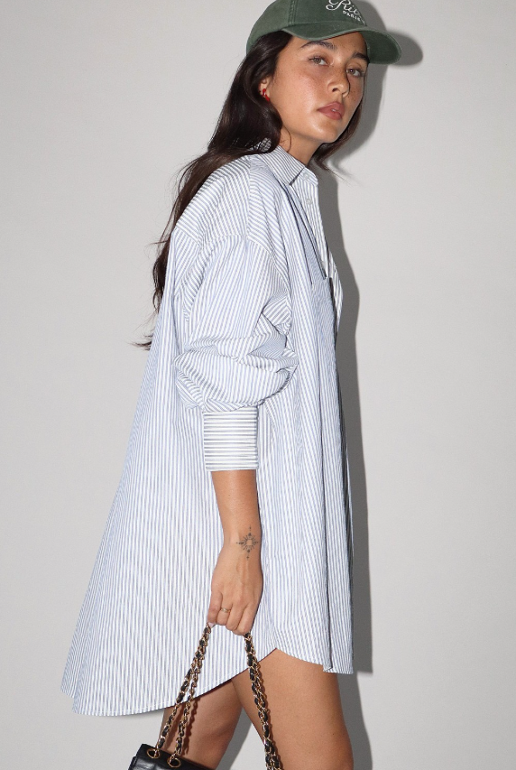 Woman wearing a long blue and white striped shirt dress, holding a bag against a plain background