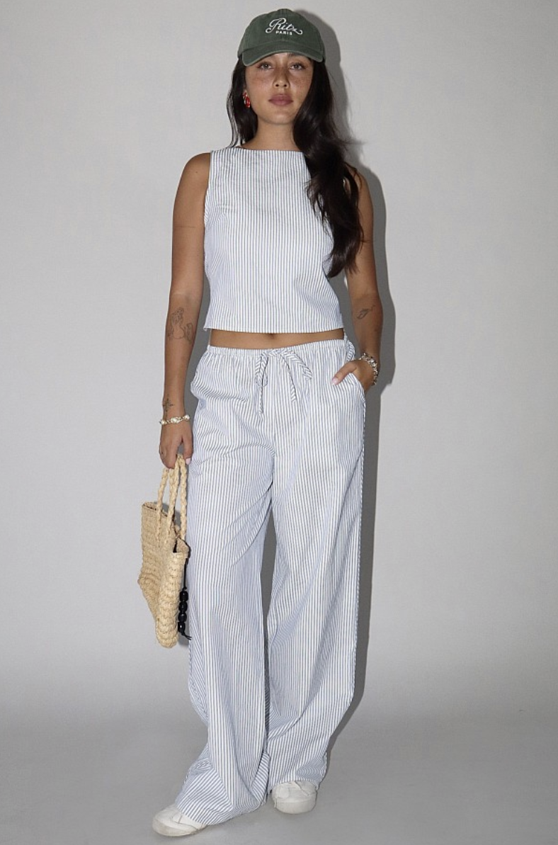 A woman wearing a striped sleeveless top and matching striped pants, holding a woven bag, on a light background.