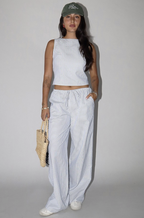 A woman wearing a striped sleeveless top and matching striped pants, holding a woven bag, on a light background.