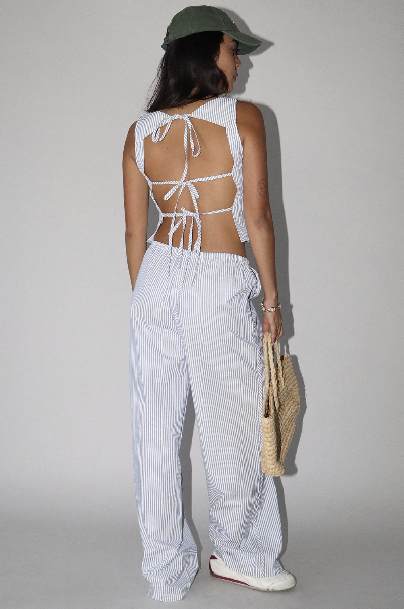 A woman wearing a striped, tie back tank top and matching striped pants and a green hat holding a woven bag on a light background.
